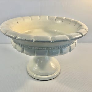 May include: A white pedestal planter with a scalloped rim and a round base. The planter has a light blue accent color and a cream-colored interior. The planter is made of a solid material and is suitable for indoor or outdoor use.