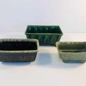 以下が含まれることがあります： 緑色の異なる色合いの長方形のセラミックプランターが3つあります。最大のプランターは幾何学的なパターンがあり、真ん中のプランターは濃い緑色の釉薬がかかっており、最小のプランターは斑点のある緑色の釉薬がかかっています。