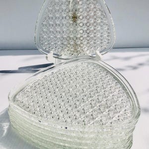 May include: A stack of clear, triangular glass plates with a textured, starburst pattern. One plate is propped up on a white stand, showcasing the design. The plates are likely for serving appetizers or desserts.