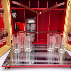 May include: A portable bar set in a tan case with a red interior, featuring a silver cocktail shaker, a metal muddler, and multiple clear glass tumblers held by brown straps. The base is a reflective surface with horizontal silver stripes.