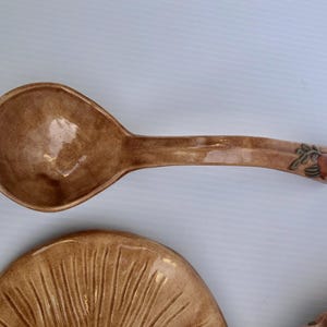 May include: A brown ceramic spoon with a wide bowl and long handle, decorated with a floral design at the end. The spoon is part of a matching set, with a plate visible below. The set has a glossy finish.