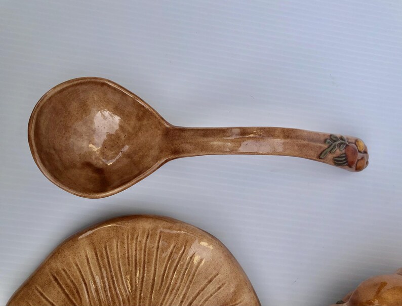 May include: A brown ceramic spoon with a wide bowl and long handle, decorated with a floral design at the end. The spoon is part of a matching set, with a plate visible below. The set has a glossy finish.