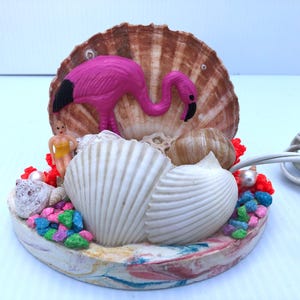 May include: A pink flamingo figurine with black eyes and beak sits on a bed of seashells and colorful pebbles. The base is made of a light brown material with a marbled effect. A white cord is attached to the base.