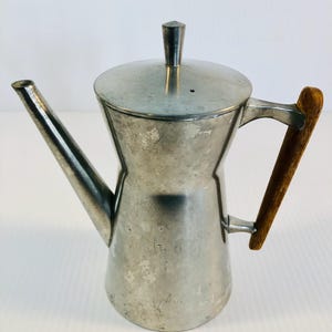 Puede incluir: Una cafetera vintage de color plateado con un asa de madera. La cafetera tiene un pico largo y una tapa con un pequeño asa geométrica. El asa es de madera de color marrón claro. La cafetera es probablemente de metal.