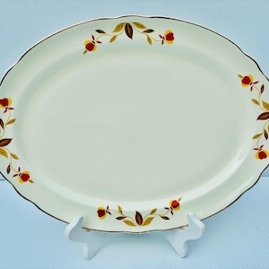 Puede incluir: Una bandeja de servir ovalada de cerámica color crema con borde festoneado y acabado en tono dorado. La bandeja presenta un patrón floral repetitivo de pequeñas flores rojas y amarillas con hojas marrones dispuestas a lo largo del borde.