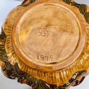 May include: A vintage ceramic bowl in shades of brown and green. The base is stamped with a logo and the year "1979". The rim of the bowl has a textured, leaf-like pattern.