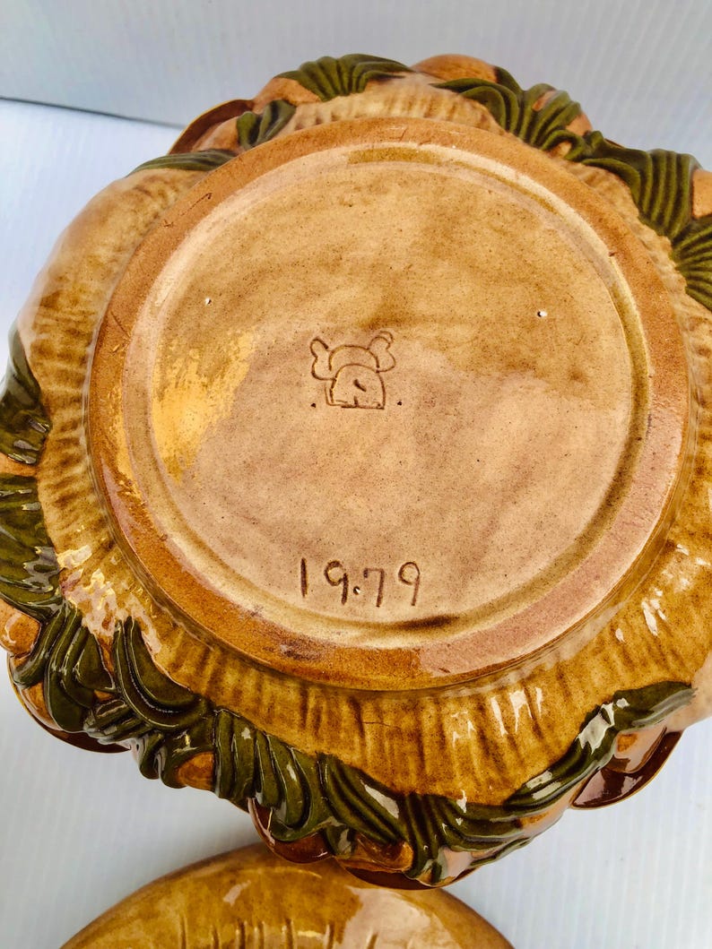 May include: A vintage ceramic bowl in shades of brown and green. The base is stamped with a logo and the year "1979". The rim of the bowl has a textured, leaf-like pattern.