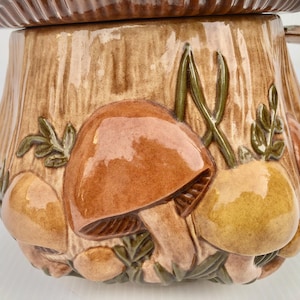 May include: A ceramic mushroom-shaped container with a lid. The container is brown with a textured, tree-trunk design. The container is decorated with raised, glossy mushrooms in shades of brown and yellow, with green accents.