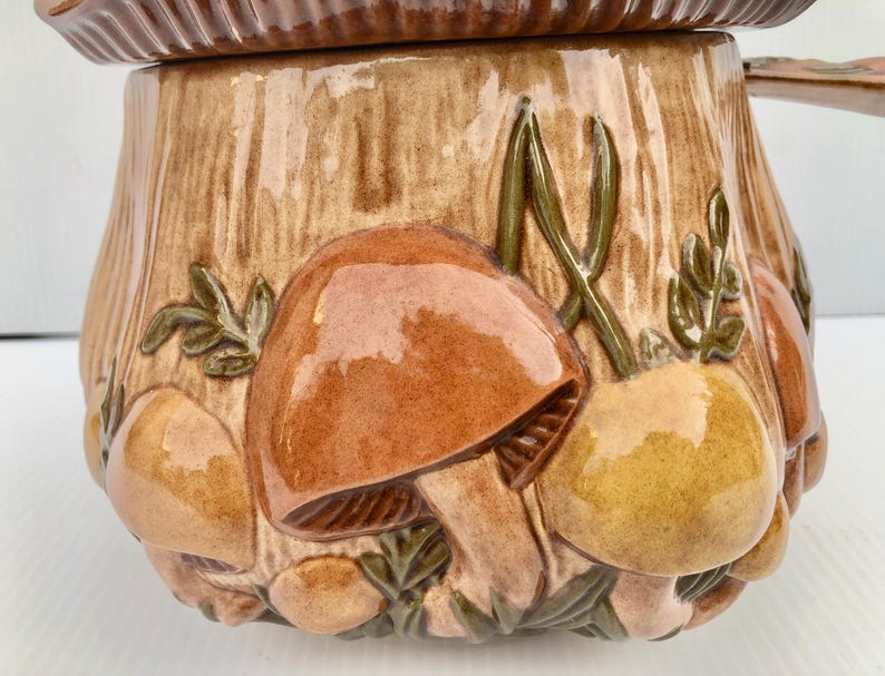May include: A ceramic mushroom-shaped container with a lid. The container is brown with a textured, tree-trunk design. The container is decorated with raised, glossy mushrooms in shades of brown and yellow, with green accents.