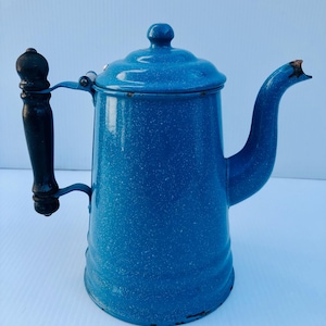 Puede incluir: Cafetera vintage de esmalte azul con asa de madera negra. La cafetera tiene un acabado moteado y una tapa con un pequeño pomo. El pico es curvo y la cafetera muestra signos de desgaste, lo que le da encanto.