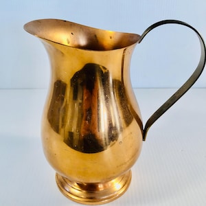 May include: A copper pitcher with a brass handle. The pitcher has a rounded body and a flared lip.