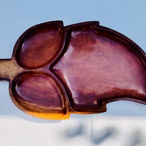 Puede incluir: Una bandeja de madera con forma de hoja con tres compartimentos y un asa. La bandeja está hecha de madera de color marrón oscuro con un acabado pulido. La bandeja está diseñada para servir aperitivos.