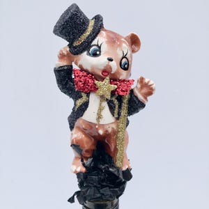 May include: A whimsical bottle stopper featuring a brown bear figurine dressed in a black tuxedo, top hat, and red bow tie. The bear holds a gold star wand. The stopper is attached to a wine bottle.