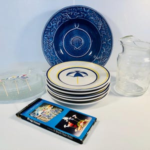 May include: A selection of beach-themed tableware and accessories. Includes a large blue plate with seashell details, a stack of plates with umbrella designs, a clear glass jug, and a box of beach chair picks.