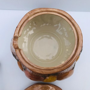 May include: A ceramic soup tureen with a lid, in brown and tan tones. The tureen has a round shape with a handle. The interior is a light beige colour. A matching ladle is visible.