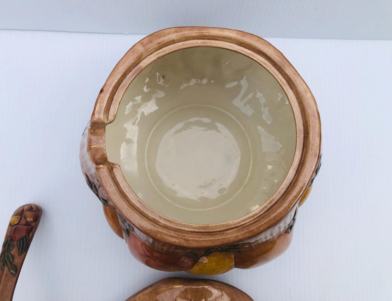 May include: A ceramic soup tureen with a lid, in brown and tan tones. The tureen has a round shape with a handle. The interior is a light beige colour. A matching ladle is visible.