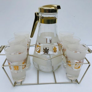May include: A set of six frosted glass tumblers and a pitcher with gold leaf accents in a gold wire rack. The glasses and pitcher have a white background with gold leaf squares and leaf designs.