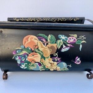 Puede incluir: Revistero de metal negro con un diseño pintado de frutas y flores. El revistero tiene dos compartimentos y decoración de volutas.