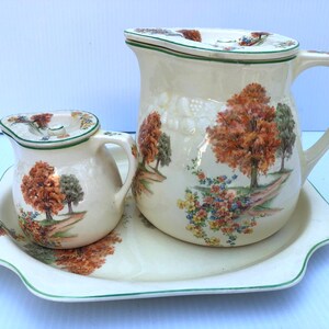 May include: A white ceramic serving tray with a green rim, featuring a cream pitcher and a smaller creamer with lids. Both pieces have a floral design with trees and flowers in shades of green, brown, and orange.