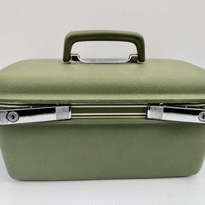 May include: Olive green vintage travel case with a textured surface, a top handle, and silver-toned latches. The case has a rectangular shape and a rounded top. A classic piece for storage.