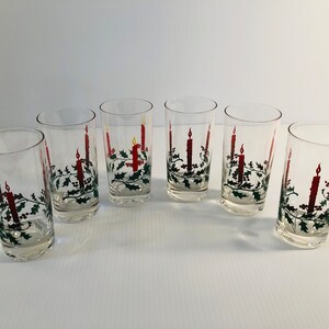 Peut inclure: Six verres transparents orn&eacute;s d'un motif de bougie de No&euml;l. Chaque verre pr&eacute;sente une bougie rouge avec une flamme jaune, entour&eacute;e de houx vert et de baies rouges. Les verres sont dispos&eacute;s sur une surface blanche.