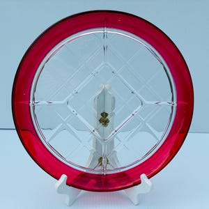 Puede incluir: Un plato redondo de cristal con un borde rojo vibrante y un centro transparente con un patrón de diamantes y cuadrícula. El plato se muestra en posición vertical sobre un soporte blanco, destacando el contraste entre el cristal y el borde.