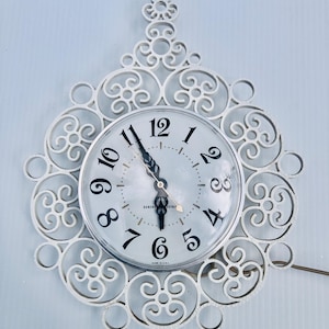 Puede incluir: Reloj de pared General Electric blanco con un marco calado decorativo. La esfera del reloj tiene números y manecillas negras, con las palabras "General Electric" impresas en la esfera. El reloj está montado en una pared azul claro.