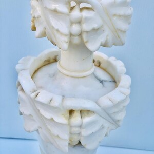 Puede incluir: Un candelabro de m&aacute;rmol blanco con intrincadas tallas de hojas y bayas. El candelabro presenta una base redonda, una columna central y dos bandas decorativas con dise&ntilde;os de hojas talladas. El dise&ntilde;o general es elegante y detallado.