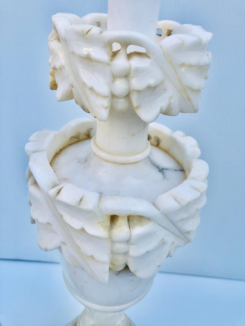 Puede incluir: Un candelabro de m&aacute;rmol blanco con intrincadas tallas de hojas y bayas. El candelabro presenta una base redonda, una columna central y dos bandas decorativas con dise&ntilde;os de hojas talladas. El dise&ntilde;o general es elegante y detallado.
