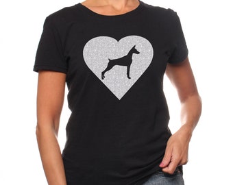 May include: A black short-sleeved t-shirt featuring a large silver glitter heart graphic on the chest. Inside the heart is a solid black silhouette of a Doberman Pinscher dog standing in profile, facing toward the right side of the garment.