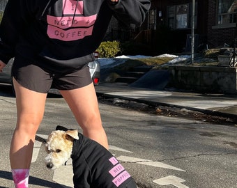 May include: A person wearing a black hoodie with pink text that says "I need a coffee" is walking with a small dog wearing a black hoodie with pink text that says "I need a puppuccino".