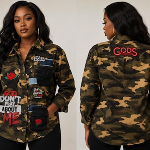 Pode incluir: Uma camisa de botões com estampa de camuflagem, patches e bordados. A frente exibe uma rosa vermelha e o texto "GOD DON'T PLAY ABOUT ME", "PRAYER CHANGES THINGS" e "God is within her she will not fall". As costas apresentam "GOD'S MASTERPIECE" em vermelho.