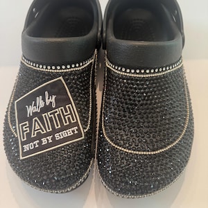 Op de afbeelding: Zwarte klompen versierd met zwarte strass-steentjes. De klompen hebben een wit en zwart label met de tekst "Walk by Faith, Not by Sight". De randen zijn afgewerkt met kleine zilveren strass-steentjes.