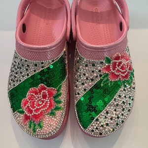 Op de afbeelding: Roze Crocs klompen versierd met strass, pailletten en geborduurde rode rozen. De klompen hebben een groene pailletten streep en parelachtige kralen. De bandjes staan omhoog.