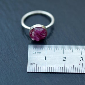 May include: A silver ring with a pink, raw ruby gemstone set in the center. The ring is shown next to a ruler measuring 4 centimeters.