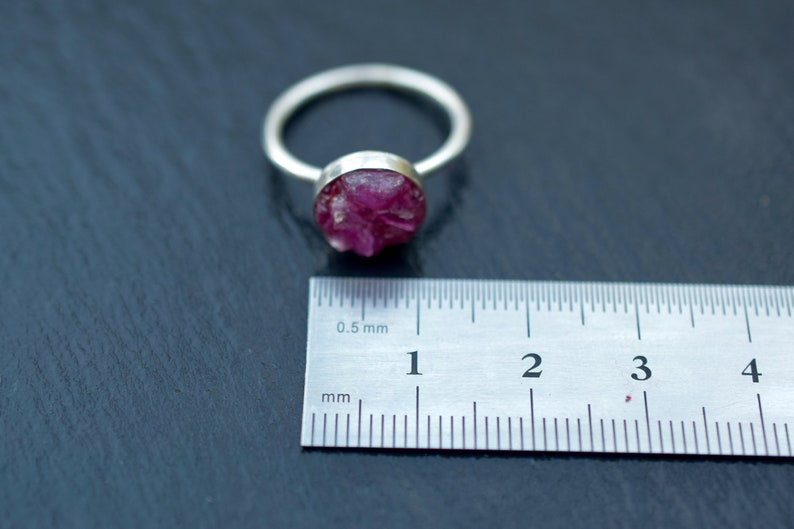 May include: A silver ring with a pink, raw ruby gemstone set in the center. The ring is shown next to a ruler measuring 4 centimeters.