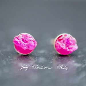 May include: A pair of silver stud earrings featuring pink ruby stones. The stones have an irregular shape and a rough, natural appearance. The text "July's Birthstone - Ruby" is written below the earrings.