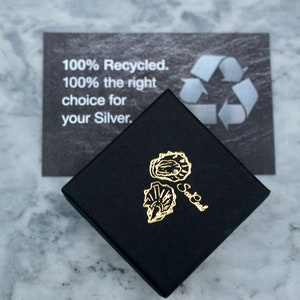 Op de afbeelding: Een zwarte vierkante doos met een gouden logo van twee oesterschelpen. De doos staat op een marmeren oppervlak. Achter de doos ligt een kaart met de tekst "100% gerecycled. 100% de juiste keuze voor uw zilver."