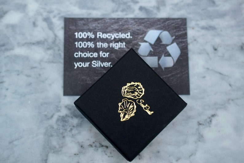 Op de afbeelding: Een zwarte vierkante doos met een gouden logo van twee oesterschelpen. De doos staat op een marmeren oppervlak. Achter de doos ligt een kaart met de tekst "100% gerecycled. 100% de juiste keuze voor uw zilver."