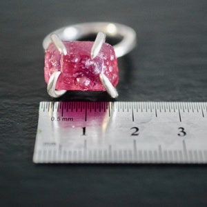 Op de afbeelding: Een zilveren ring met een roze, gefacetteerde edelsteen in een kraalbezetting. De ring wordt gemeten met een liniaal, wat laat zien dat de steen ongeveer 1 cm breed is.