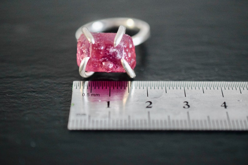 Op de afbeelding: Een zilveren ring met een roze, gefacetteerde edelsteen in een kraalbezetting. De ring wordt gemeten met een liniaal, wat laat zien dat de steen ongeveer 1 cm breed is.