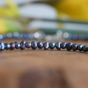 May include: A necklace of dark grey freshwater pearls. The pearls are irregularly shaped and have a lustrous sheen. The necklace is displayed on a wooden surface with a blurred background of yellow and green.