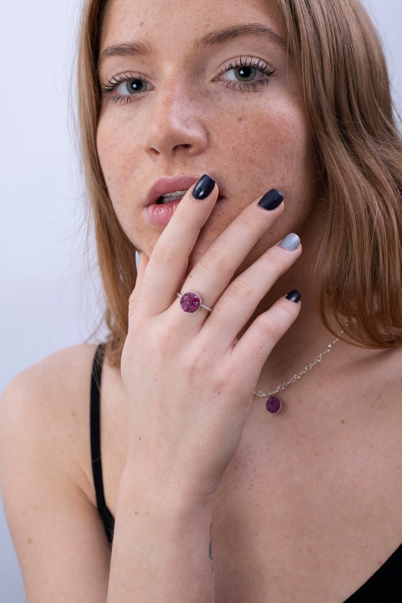 May include: A silver ring with a round, pink gemstone. The ring is on a person's finger. The person is wearing a black tank top and a silver necklace with a pink gemstone pendant.