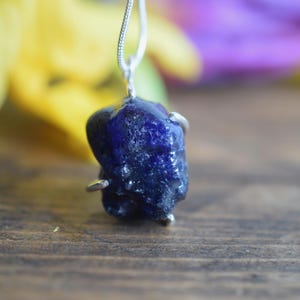 Puede incluir: Un colgante de piedra preciosa de zafiro en bruto de color azul oscuro en una cadena de plata. El zafiro es una piedra natural en bruto de color azul oscuro.