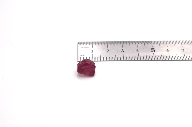 Op de afbeelding: Een dieprode edelsteen met een ruwe, onregelmatige vorm, ongeveer 1 centimeter lang. De edelsteen ligt naast een liniaal voor schaal.
