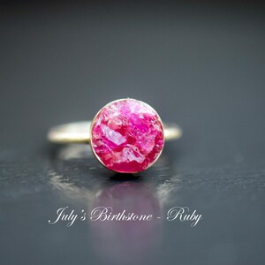 May include: A silver ring with a pink ruby gemstone. The ruby is a rough cut and has a natural, organic look. The text "July's Birthstone - Ruby" is visible below the ring.