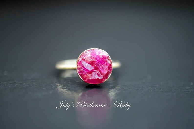May include: A silver ring with a pink ruby gemstone. The ruby is a rough cut and has a natural, organic look. The text "July's Birthstone - Ruby" is visible below the ring.