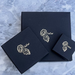 K&ouml;nnte beinhalten: Drei schwarze Boxen mit goldenen Folien-Designs von Austern und dem Text "Sea Pearl" auf jeder Box.