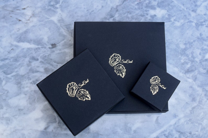 K&ouml;nnte beinhalten: Drei schwarze Boxen mit goldenen Folien-Designs von Austern und dem Text "Sea Pearl" auf jeder Box.