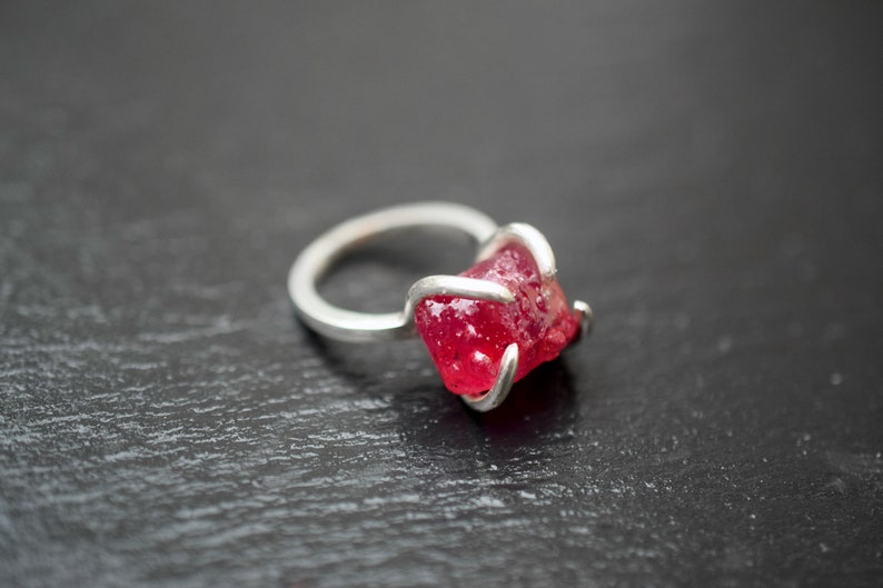 Op de afbeelding: Een zilveren ring met een grote, gefacetteerde, rode edelsteen gezet in een eenvoudig, modern ontwerp.